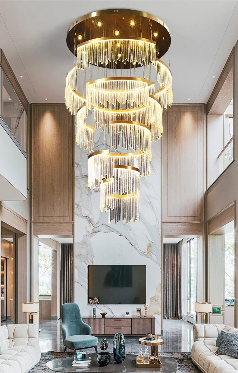 A luxurious, spacious living room featuring a large gold tiered crystal waterfall chandelier as the central light source, complemented by modern furniture and a large television.