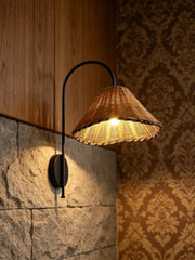 Close-up of a black metal arm wall sconce with a woven rattan cone shade against a stone wall with patterned wallpaper.