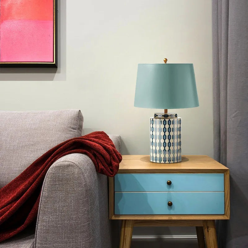 A blue ceramic table lamp with a geometric pattern and a light blue fabric shade sits on a wooden nightstand next to a grey armchair with a red throw.