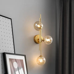 A gold wall sconce with clear glass globes showcasing illuminated warm LED bulbs, mounted on a brass stem next to framed artwork and gray curtains.