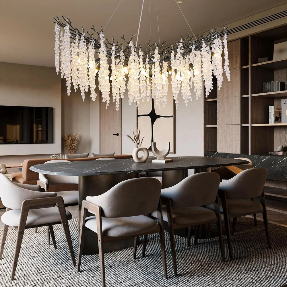 A curved, branch-like chandelier with hanging crystals, suspended above an oval dining table in a well-decorated room.