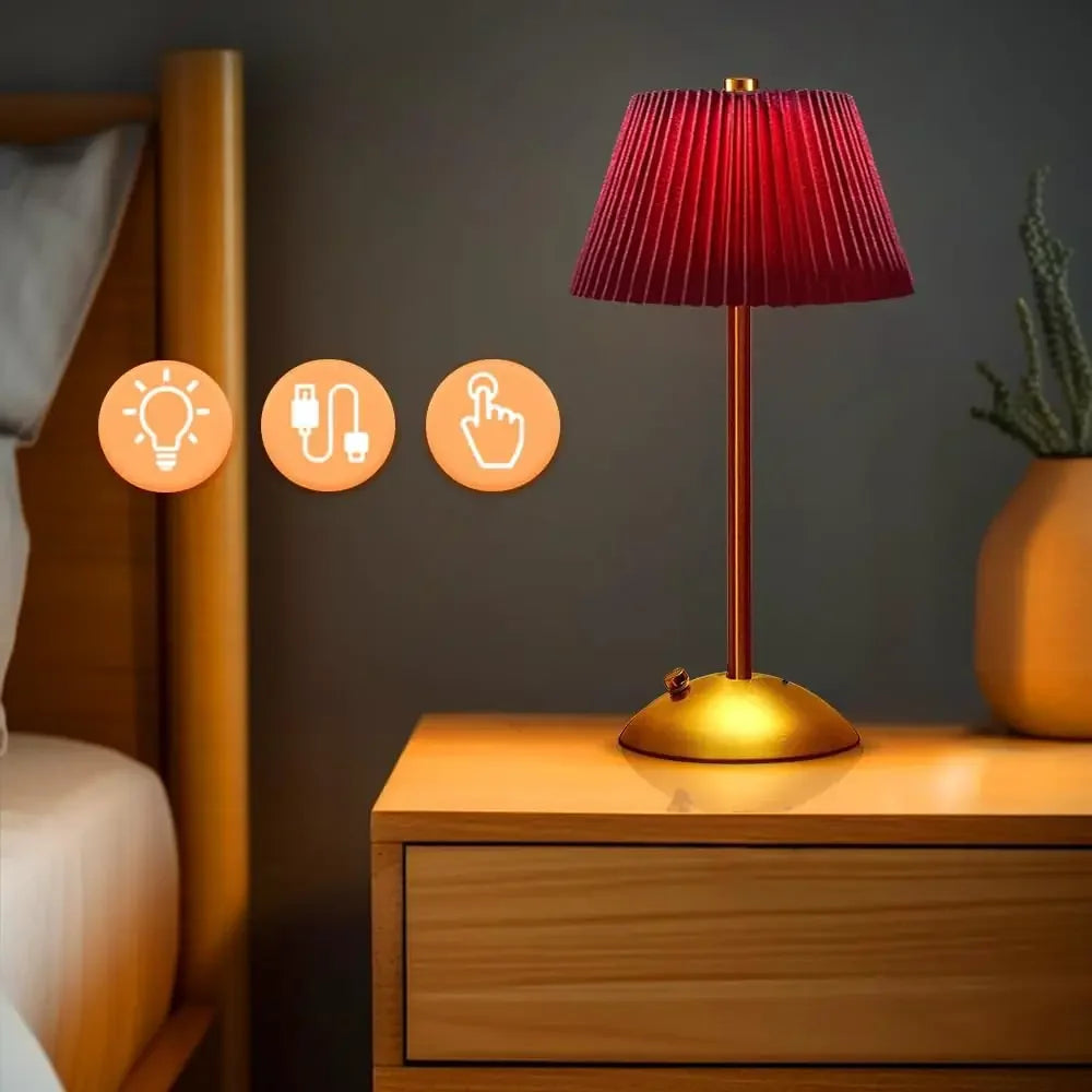A red pleated fabric shade table lamp with a gold base and stem, placed on a wooden nightstand next to a bed, with icons indicating light, USB, and touch control.