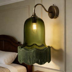 A wall-mounted brass sconce with a green bell-shaped glass shade and a fringe of green tassels, casting a warm glow on a wall next to a bed with white linens.