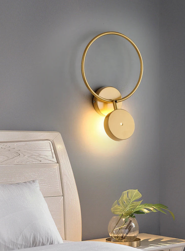 Gold wall sconce on a gray wall with a bed and plant in the foreground