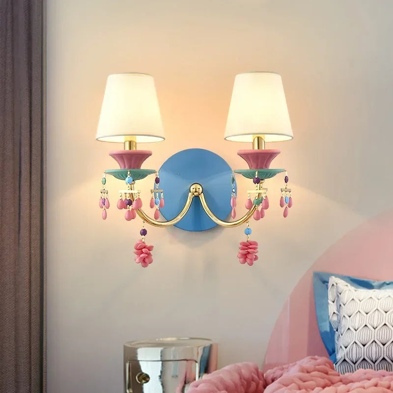 Gold wall sconce with white fabric shades and colorful beaded and flower-like accents, mounted on a blue backing plate, shown in a room setting.