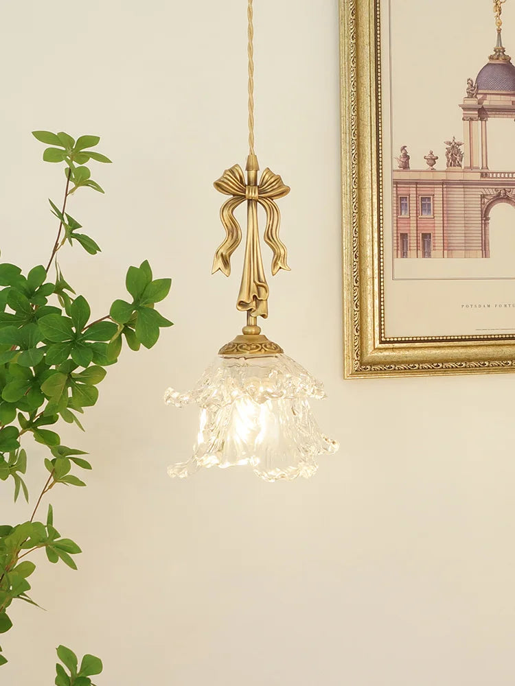 Decorative gold pendant light with clear glass shade on a neutral wall with a plant and framed picture.