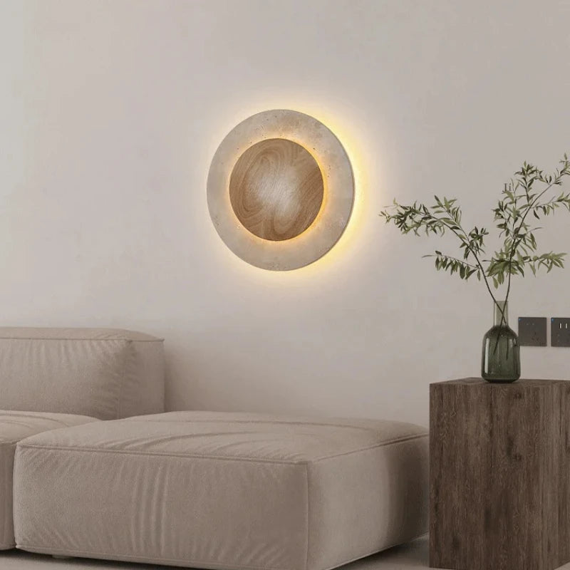 A contemporary round stone wall light with a warm LED glow, positioned on a white wall above a beige upholstered ottoman, with a wooden side table holding a vase of greenery.