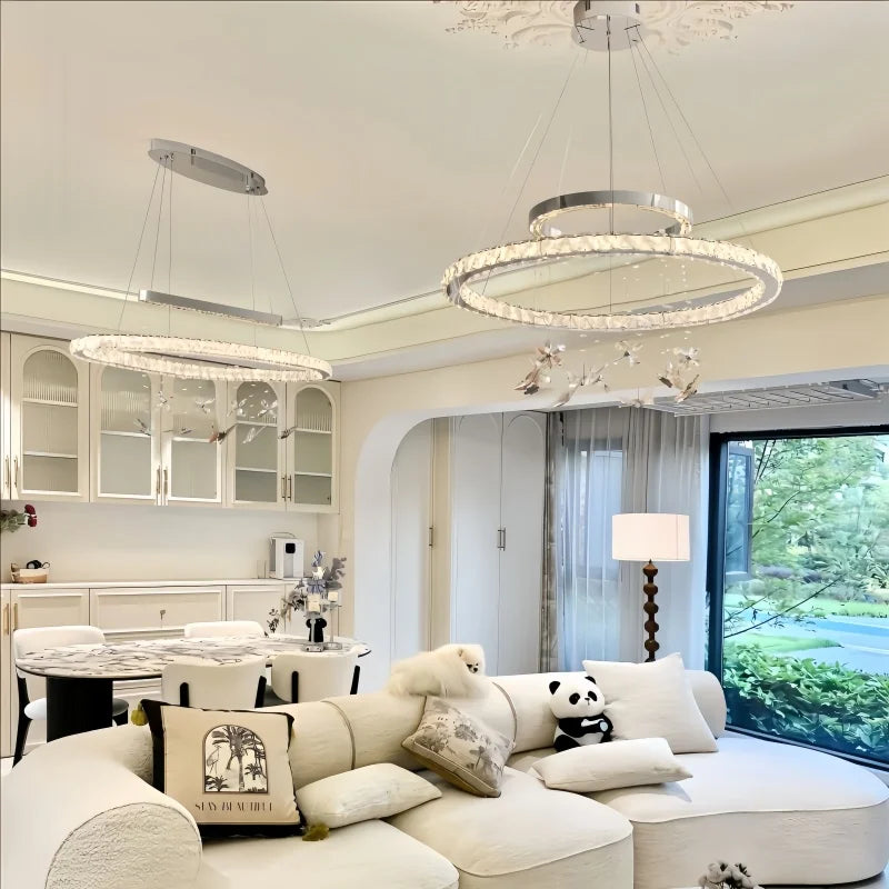 A spacious living room with two distinct chandeliers: a circular multi-tiered crystal chandelier and a long, oval crystal chandelier, both featuring hanging butterfly ornaments.