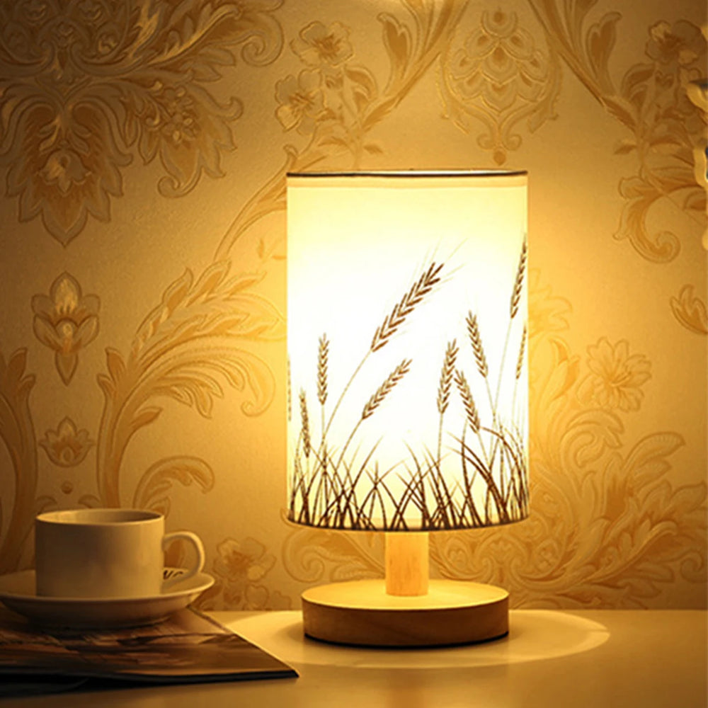 Linen drum shade table lamp with a subtle wheat stalk pattern on a wooden base, casting a warm glow next to a cup and saucer.