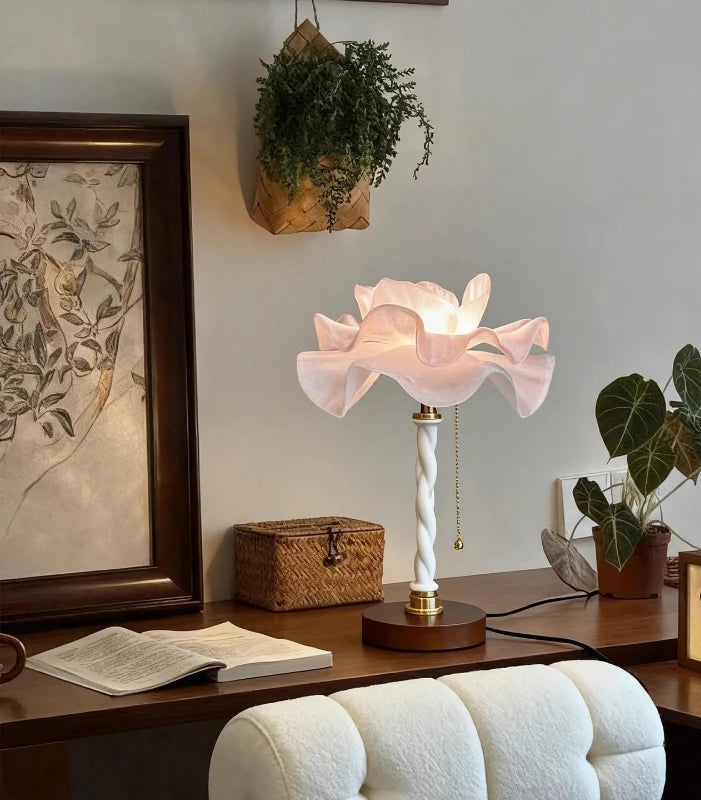 A white and pink petal-shaped table lamp sitting on a wooden desk next to an open book and a small woven basket.