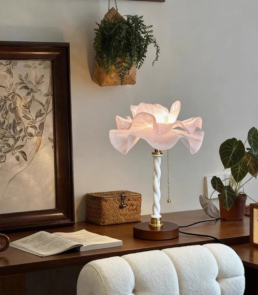 A white and pink petal-shaped table lamp sitting on a wooden desk next to an open book and a small woven basket.