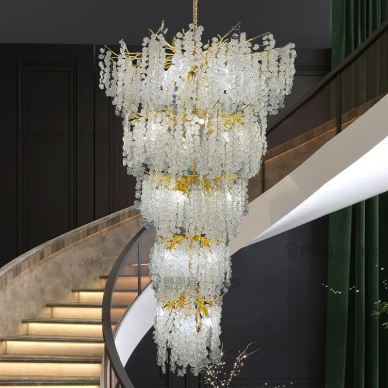 A grand, multi-tiered gold chandelier with cascading clear crystals hangs in a modern interior with a curved staircase.