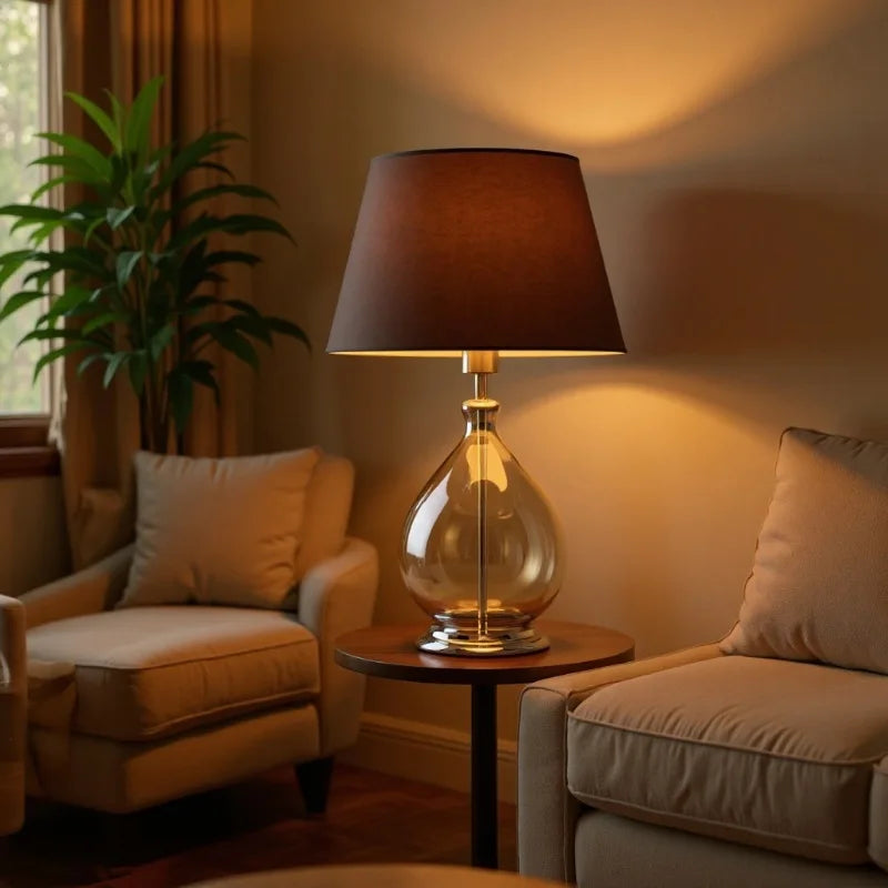 Amber glass table lamp with dark fabric shade on a small side table between two armchairs, with a plant visible through a window.