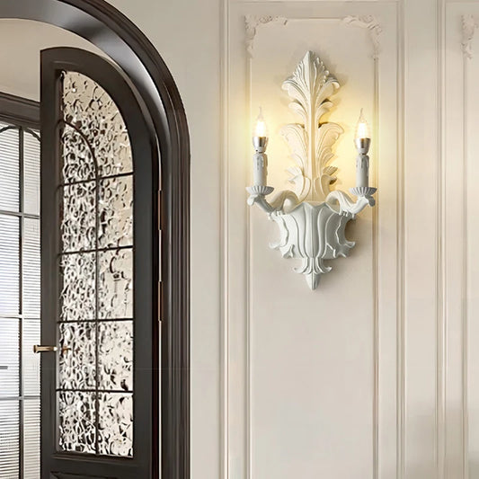 White leaf-shaped wall sconce with two candle-style lights mounted on a paneled wall, next to an arched doorway.