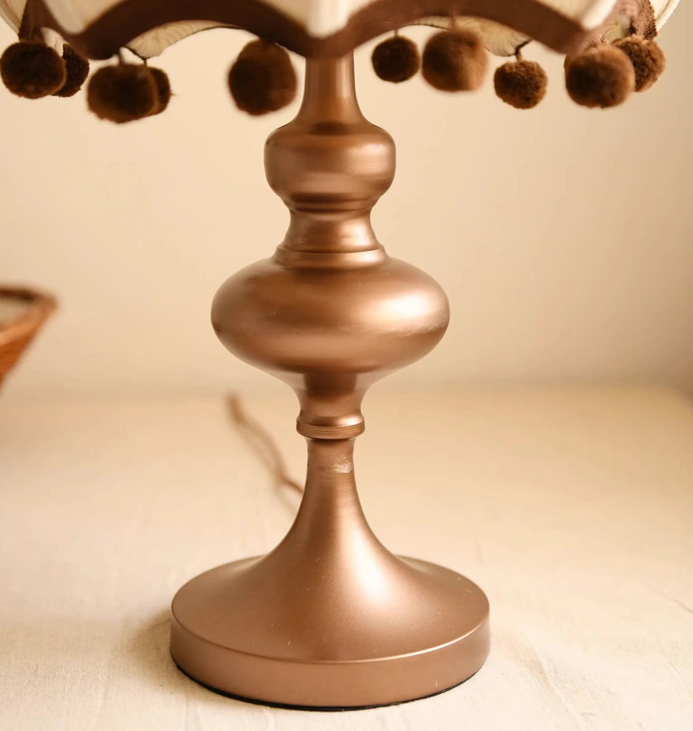 Bronze-colored decorative lamp with pom-pom details on a beige surface