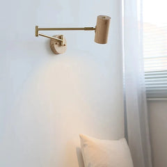 A wall sconce with a light wood cylinder lamp head and a brass swing arm mounted on a round wooden base against a white wall, illuminated by soft light.