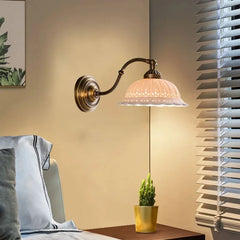 A gold and white ceramic floral wall sconce illuminated above a bedside table with a small potted plant.