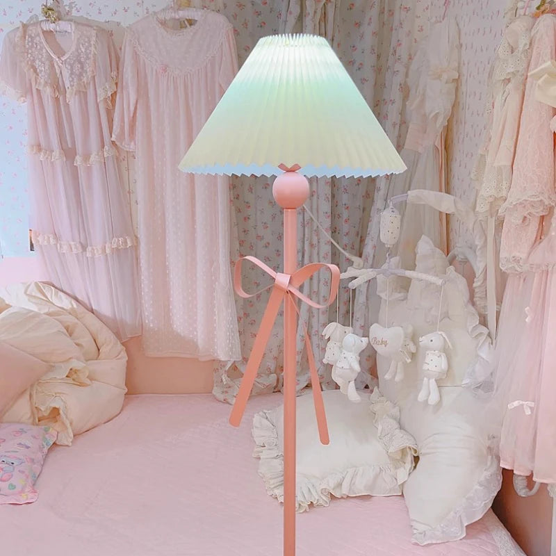 A pink floor lamp with a pleated shade and bow, placed in a room with floral wallpaper and soft pink bedding.