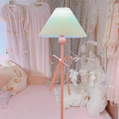 A pink floor lamp with a pleated shade and bow, placed in a room with floral wallpaper and soft pink bedding.