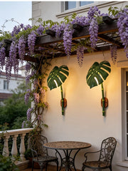 Two green leaf bamboo wall sconces mounted on an exterior wall under a blooming wisteria vine, above a small outdoor table and chairs.