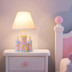 A pink castle table lamp with a conical off-white fabric shade rests on a white bedside table beside a pink bed frame.