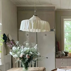 White pleated fabric pendant light hanging over a wooden dining table with a large floral arrangement.