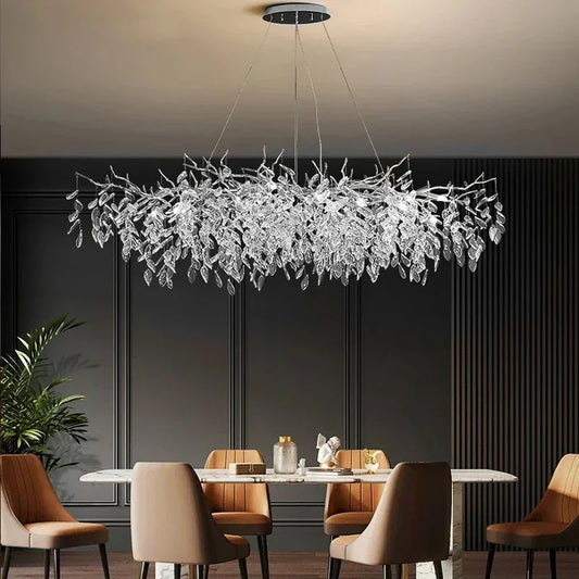 A long, horizontal chandelier with a gold frame and numerous clear crystal leaf-like droplets hanging from branches, suspended over a dining table.
