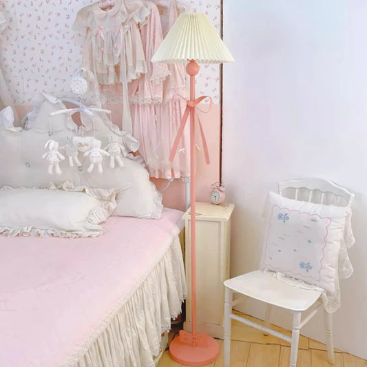 Pink floor lamp with a pleated fabric shade and a large pink bow accent, standing next to a bed with ruffled bedding.