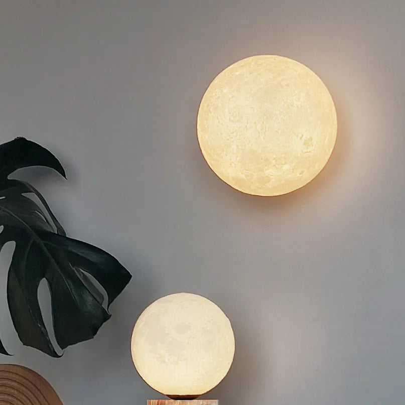 Two 3D printed moon shaped lights are displayed on a gray wall. The larger one is a wall sconce, and a smaller one sits on a wooden surface below. A large green leaf plant is in the left foreground.