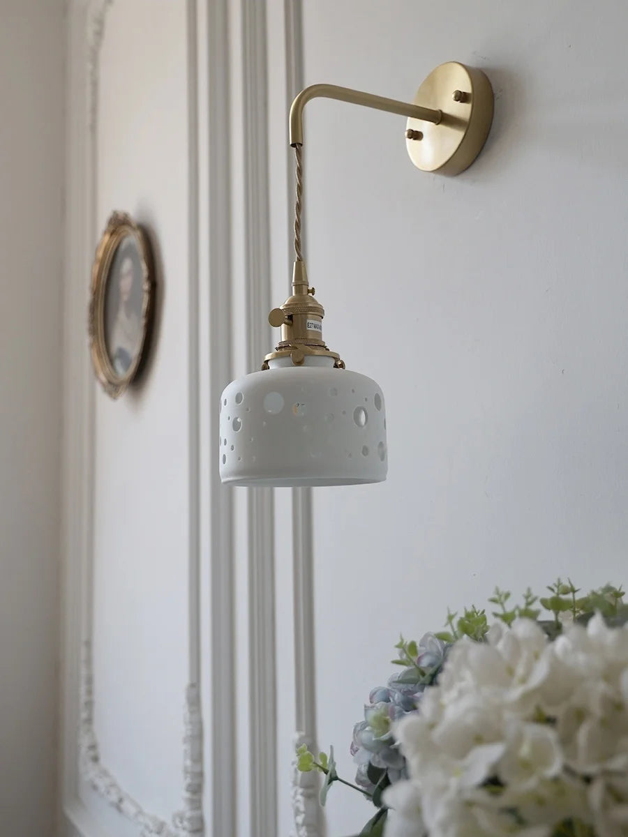Wall-mounted lamp with a decorative white shade and gold arm, featuring a clock and flowers in the background.