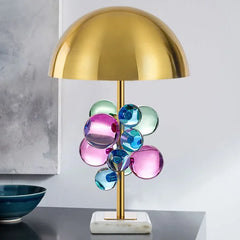 A modern table lamp with a brass finish stem, colorful glass orbs, and a white marble base, set against a neutral background.