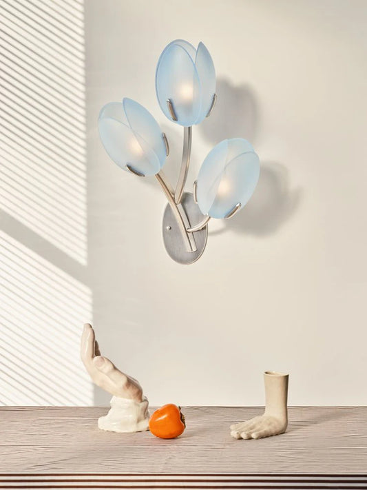 Gold floral bud wall sconce with three frosted glass shades emitting a soft blue light, mounted on a neutral wall.