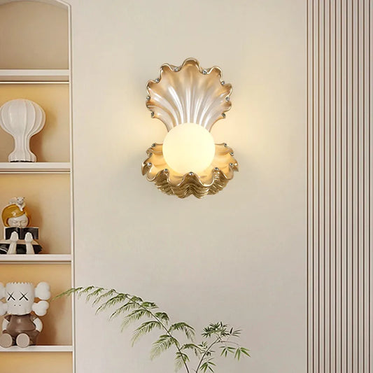 Gold seashell wall sconce with a frosted globe light, mounted on a cream-colored wall next to a bookshelf.