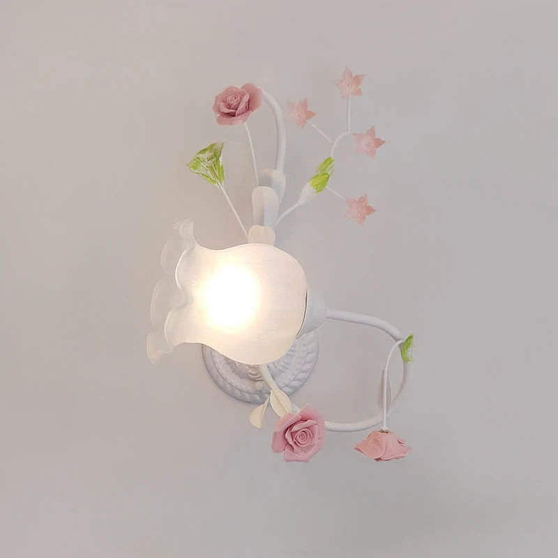 Close-up of a white floral rose bud wall sconce with a frosted glass shade illuminated, showcasing detailed pink roses and green leaves.
