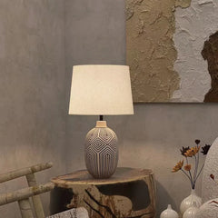 Close-up of a ceramic table lamp with a distinctive geometric pattern on its base, topped with a cream fabric shade, on a textured wooden surface.