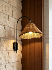 Black metal arm wall sconce with a woven rattan cone shade mounted on a stone wall next to a wooden panel.