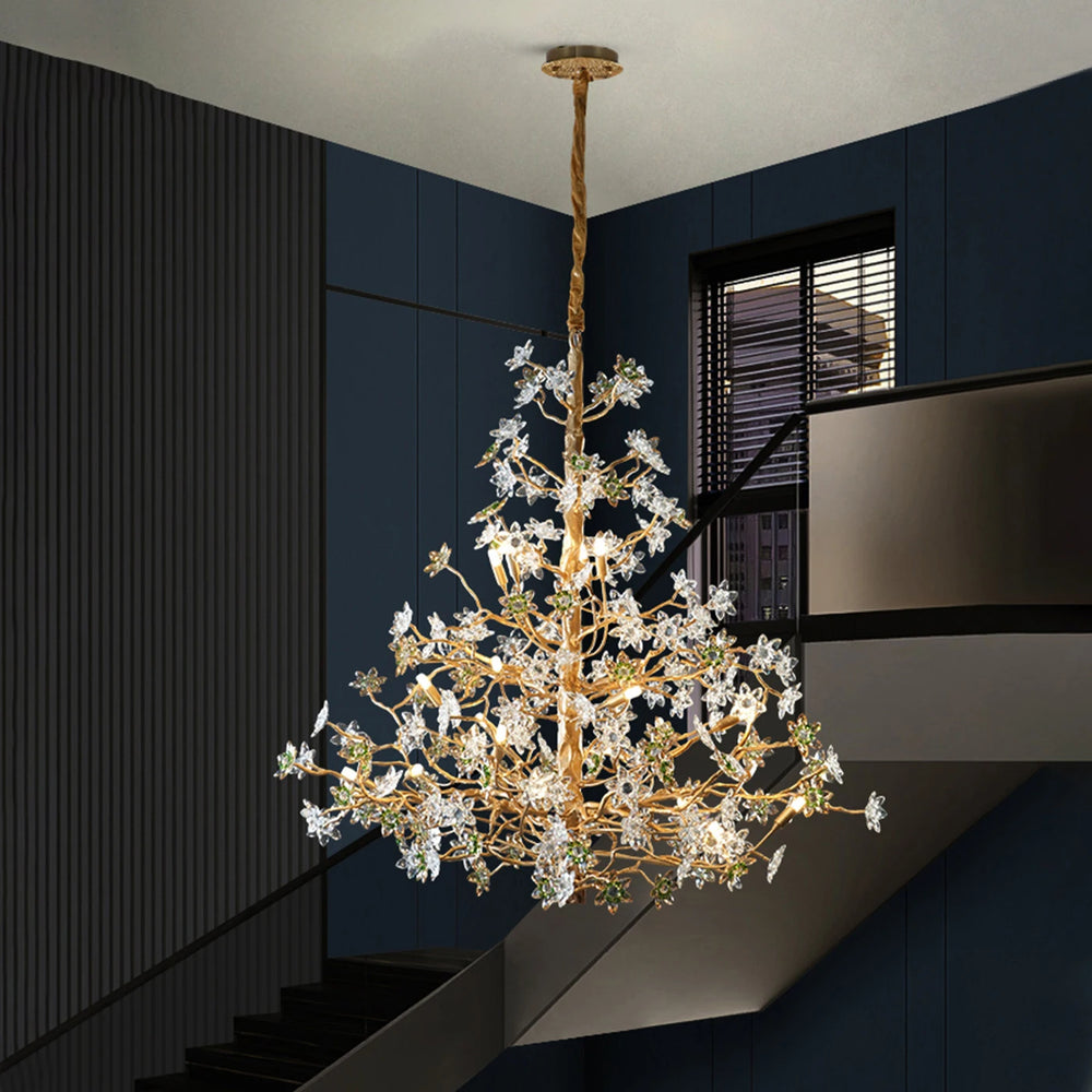 A close-up view of a gold chandelier with intricate branches adorned with clear and green crystal floral elements, suspended against a dark blue wall.