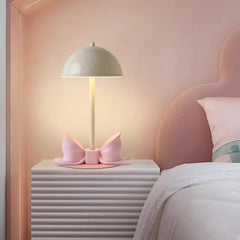 Pink bow accent table lamp with a white dome shade on a ribbed nightstand next to a pink headboard.