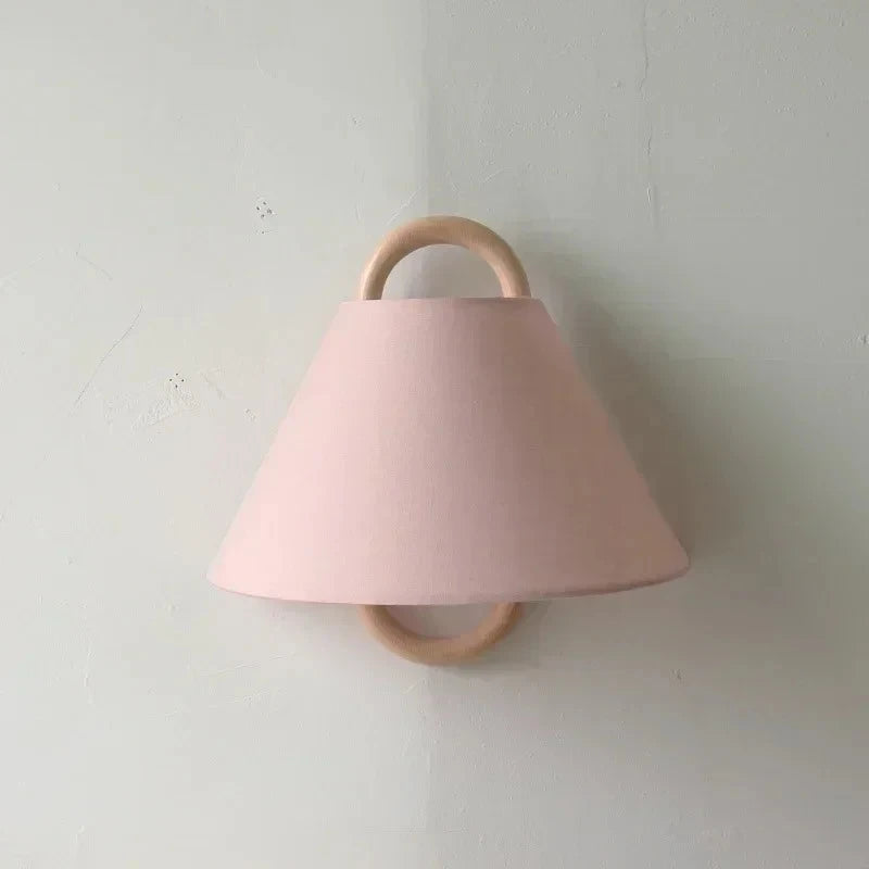 A pink fabric cone shade wall sconce with a natural wood hoop frame mounted on a textured white wall.