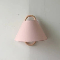A pink fabric cone shade wall sconce with a natural wood hoop frame mounted on a textured white wall.