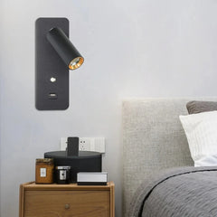 A black wall sconce with a gold spotlight and USB port, mounted on a wall above a wooden nightstand.