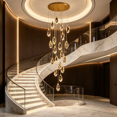 Grand foyer with a large, cascading gold chandelier featuring numerous teardrop-shaped faceted crystals hanging from a round gold ceiling plate, illuminating a marble spiral staircase.