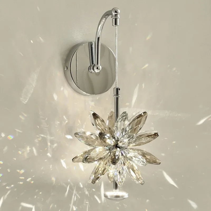 Close-up of a wall-mounted light fixture with a faceted crystal flower design in a warm tone, mounted on a polished chrome arm, casting prismatic light.