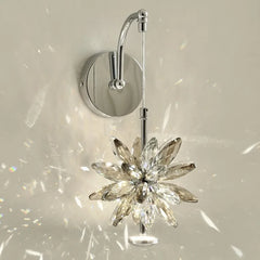 Close-up of a wall-mounted light fixture with a faceted crystal flower design in a warm tone, mounted on a polished chrome arm, casting prismatic light.