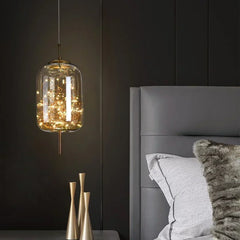 A glass cylinder pendant light filled with warm LED lights, hanging next to a modern bed.