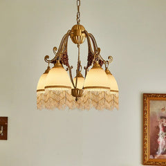 A five-light polished brass chandelier with white textured glass shades and beaded fringe, viewed from the side with a framed painting in the background.