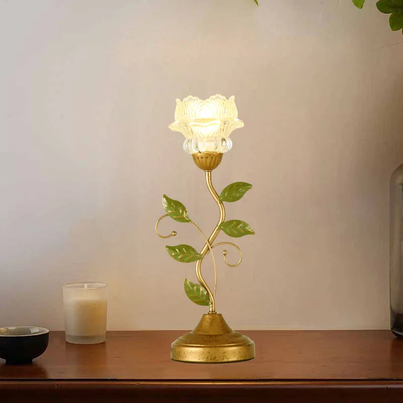 A gold table lamp with a flower-shaped glass shade and green leaves on a stem, set against a neutral background.