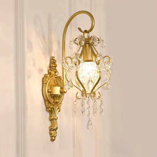 A gold wall sconce with clear crystal accents mounted on a white paneled wall, showcasing its ornate design and upward-facing light.