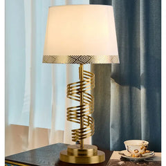 A gold spiral table lamp with a white conical fabric shade illuminated next to blue curtains.