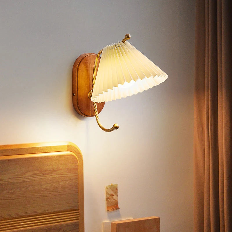 A wall sconce with a cream pleated fabric shade and gold arm mounted on a wooden base, illuminated next to a bed with wooden headboard.
